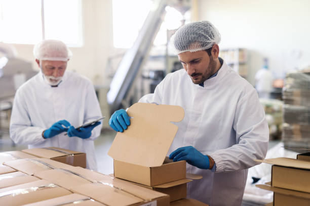 Young Caucasian employee in sterile uniform packing goods in boxes. In background supervisor holding tablet and counting boxes.